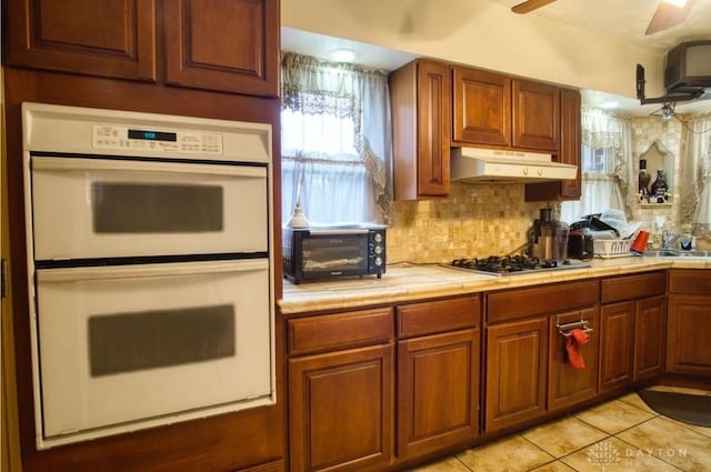 kitchen with tile counters, decorative backsplash, under cabinet range hood, double oven, and stainless steel gas stovetop