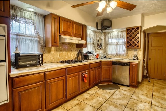 kitchen with tile counters, decorative backsplash, brown cabinetry, appliances with stainless steel finishes, and under cabinet range hood