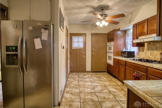 kitchen with tasteful backsplash, appliances with stainless steel finishes, brown cabinetry, light tile patterned flooring, and under cabinet range hood