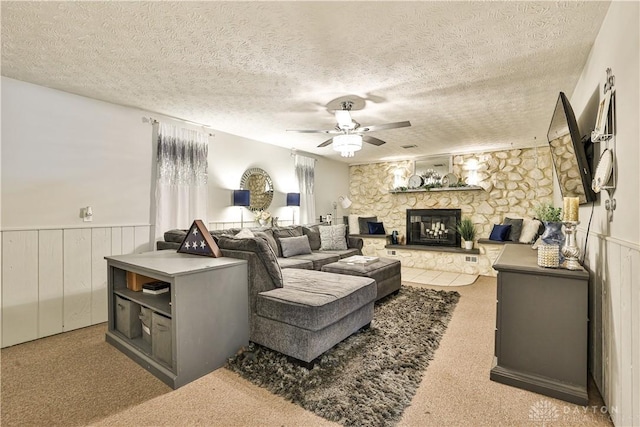 living room with a ceiling fan, a stone fireplace, a textured ceiling, wainscoting, and carpet flooring