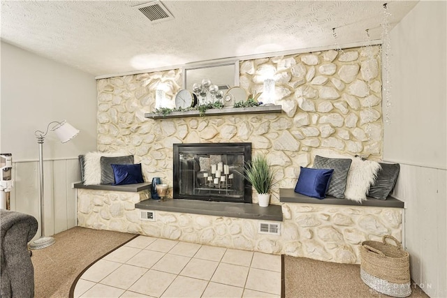 carpeted living room with a stone fireplace, a textured ceiling, wainscoting, and visible vents