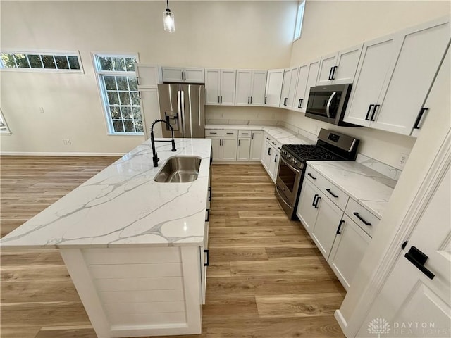 kitchen with light wood finished floors, a towering ceiling, appliances with stainless steel finishes, a kitchen island with sink, and a sink