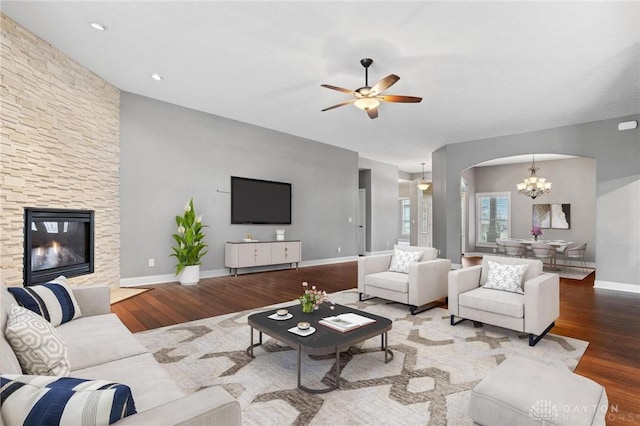 living room with baseboards, arched walkways, wood finished floors, a fireplace, and ceiling fan with notable chandelier