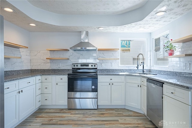 kitchen featuring stainless steel appliances, a sink, wall chimney range hood, light wood-type flooring, and open shelves