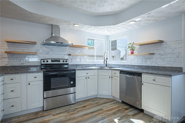 kitchen with a sink, appliances with stainless steel finishes, wall chimney range hood, open shelves, and light wood finished floors