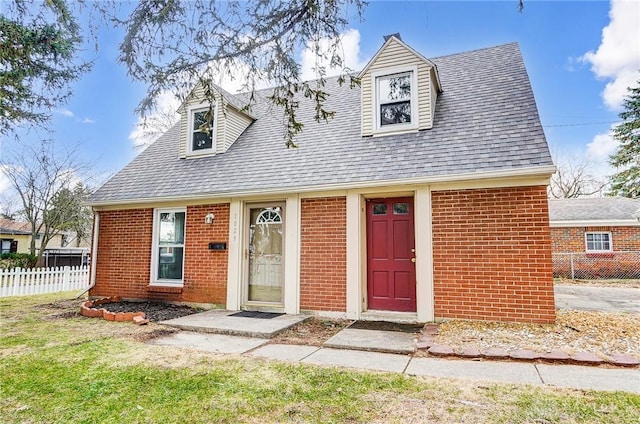 new england style home featuring a shingled roof, fence, and brick siding