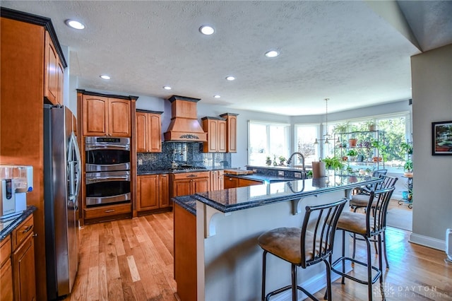 kitchen with premium range hood, a sink, a kitchen breakfast bar, appliances with stainless steel finishes, and brown cabinets