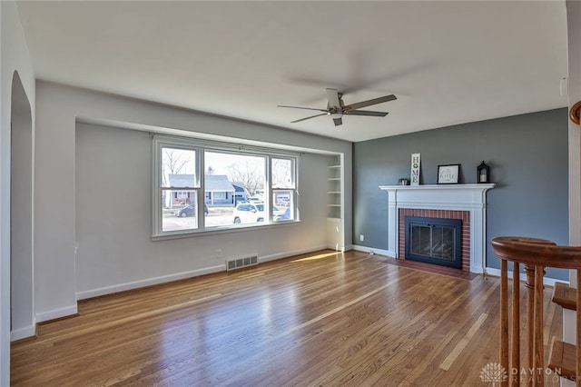 unfurnished living room featuring wood finished floors, a ceiling fan, visible vents, baseboards, and a brick fireplace