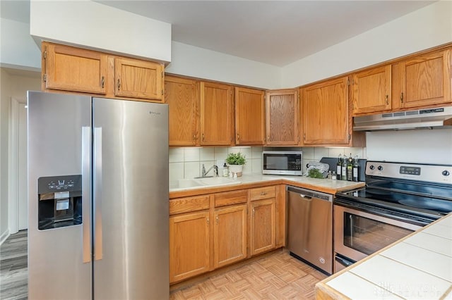 kitchen with under cabinet range hood, stainless steel appliances, a sink, tile counters, and tasteful backsplash