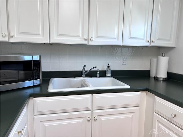 kitchen with white cabinets, dark countertops, stainless steel microwave, a sink, and backsplash