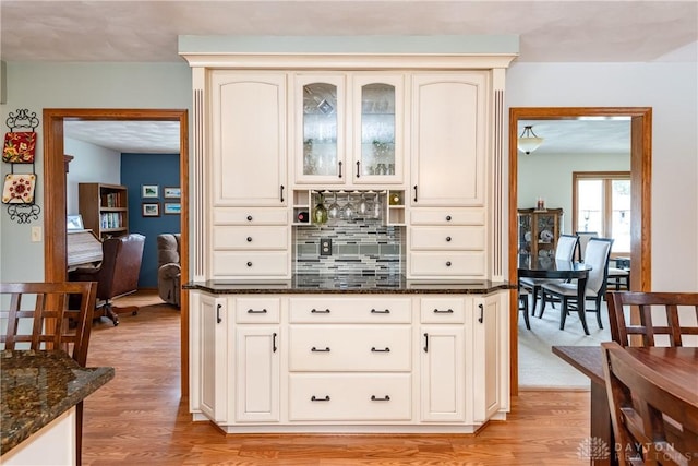 kitchen featuring dark stone counters, glass insert cabinets, cream cabinets, light wood-style floors, and backsplash