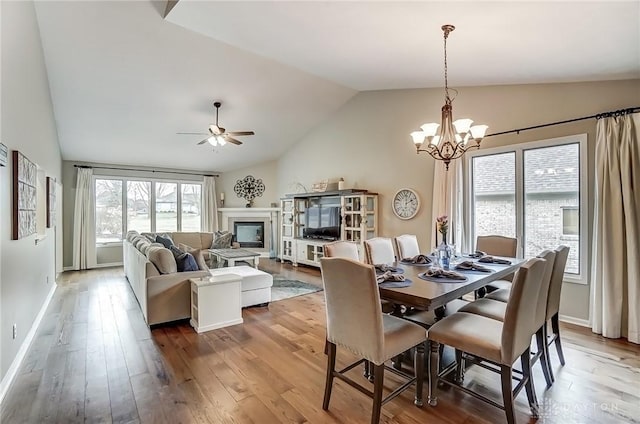 dining area featuring baseboards, a glass covered fireplace, lofted ceiling, wood finished floors, and ceiling fan with notable chandelier