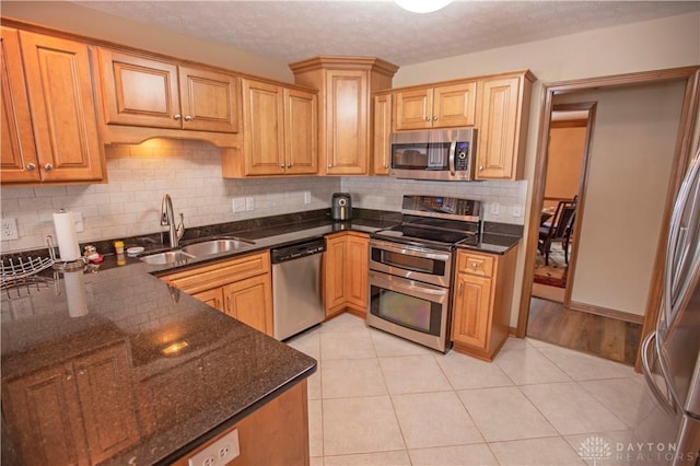 kitchen featuring light tile patterned floors, a sink, appliances with stainless steel finishes, backsplash, and dark stone counters