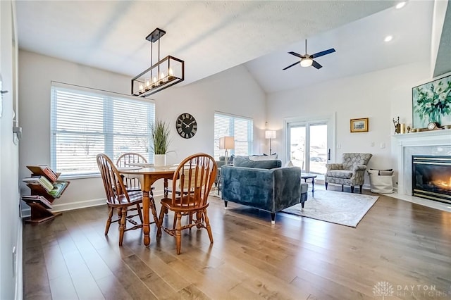 dining room with lofted ceiling, a fireplace with flush hearth, wood finished floors, baseboards, and ceiling fan with notable chandelier