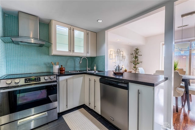 kitchen featuring dark countertops, appliances with stainless steel finishes, a sink, a peninsula, and wall chimney exhaust hood