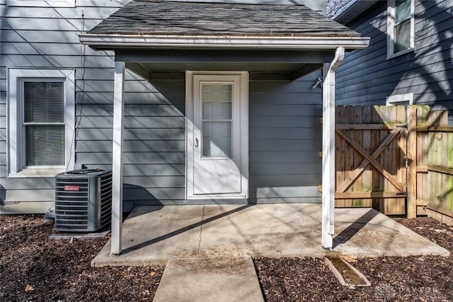 view of exterior entry featuring fence, cooling unit, and roof with shingles