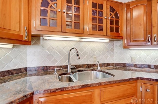 kitchen featuring tasteful backsplash, brown cabinetry, dark countertops, glass insert cabinets, and a sink