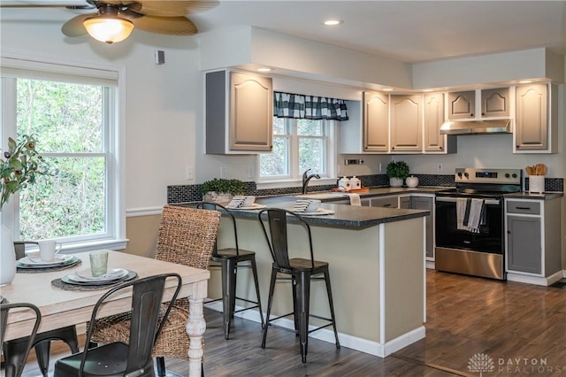 kitchen with dark countertops, a breakfast bar area, a peninsula, stainless steel electric stove, and under cabinet range hood