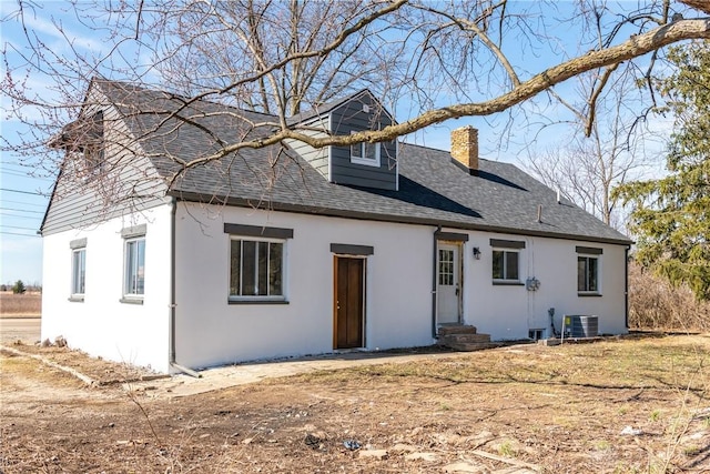 view of front facade with entry steps, cooling unit, roof with shingles, stucco siding, and a chimney