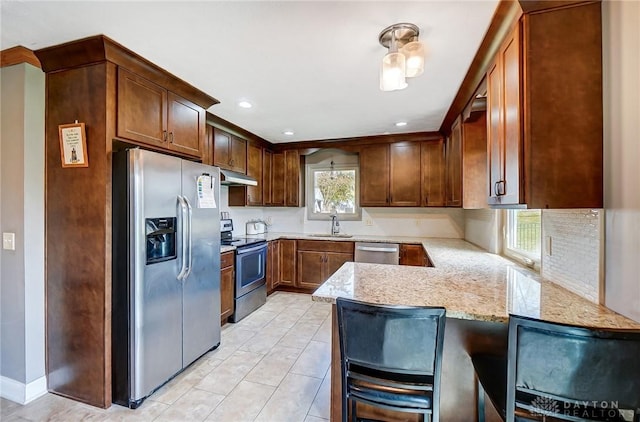 kitchen with appliances with stainless steel finishes, a sink, a peninsula, light stone countertops, and under cabinet range hood