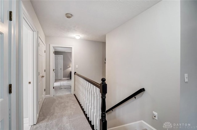 hallway featuring baseboards, light colored carpet, a textured ceiling, and an upstairs landing