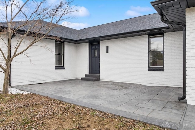 view of exterior entry featuring a shingled roof, brick siding, and a patio