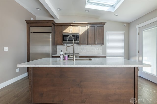 kitchen featuring tasteful backsplash, vaulted ceiling with skylight, an island with sink, light stone counters, and stainless steel appliances