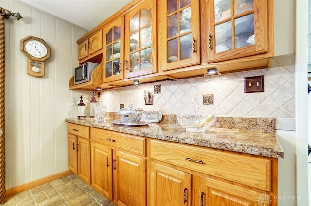 kitchen with tasteful backsplash, brown cabinetry, glass insert cabinets, light stone counters, and stainless steel microwave