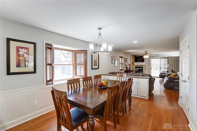 dining area with a fireplace, a decorative wall, wainscoting, wood finished floors, and ceiling fan with notable chandelier