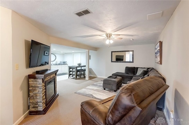 living room with light colored carpet, visible vents, ceiling fan, a stone fireplace, and a textured ceiling