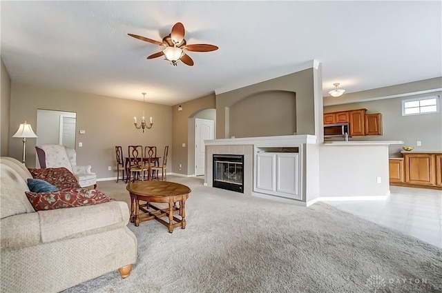 living room featuring baseboards, ceiling fan with notable chandelier, a tiled fireplace, and light colored carpet