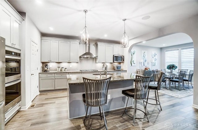 kitchen with arched walkways, stainless steel appliances, wall chimney range hood, and a sink