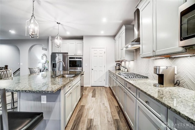 kitchen featuring a sink, light wood-style floors, appliances with stainless steel finishes, decorative backsplash, and wall chimney exhaust hood