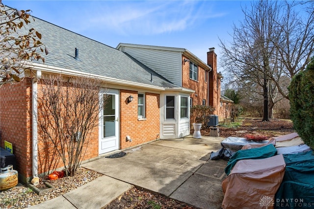 back of property with a chimney, roof with shingles, cooling unit, a patio area, and brick siding