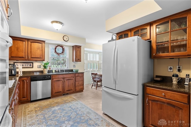 kitchen featuring light tile patterned floors, brown cabinetry, glass insert cabinets, freestanding refrigerator, and stainless steel dishwasher