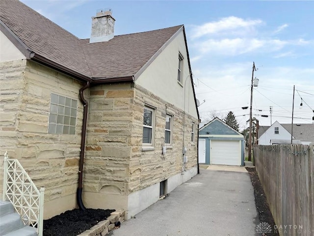 view of side of property with an outbuilding, a detached garage, fence, driveway, and roof with shingles