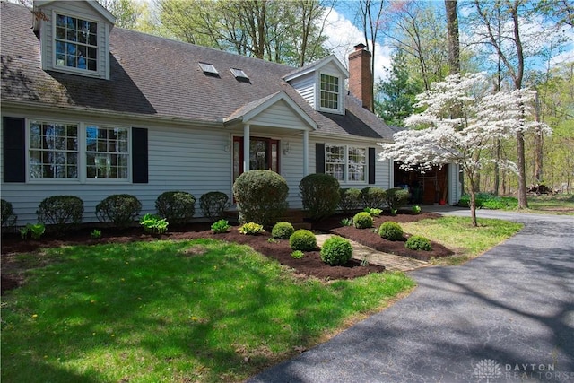 new england style home featuring driveway, a chimney, a front lawn, and roof with shingles