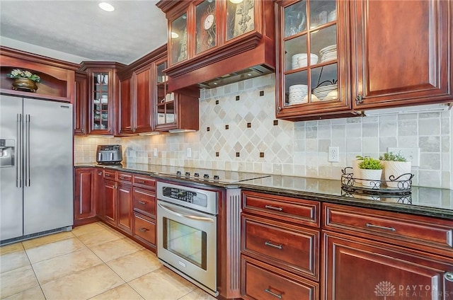 kitchen with reddish brown cabinets, light tile patterned floors, tasteful backsplash, dark stone countertops, and stainless steel appliances