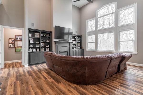 living room featuring a ceiling fan, a glass covered fireplace, wood finished floors, baseboards, and a towering ceiling