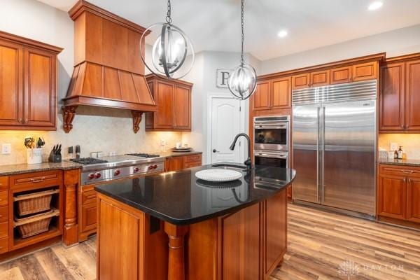 kitchen featuring light wood-type flooring, custom range hood, brown cabinets, appliances with stainless steel finishes, and a sink