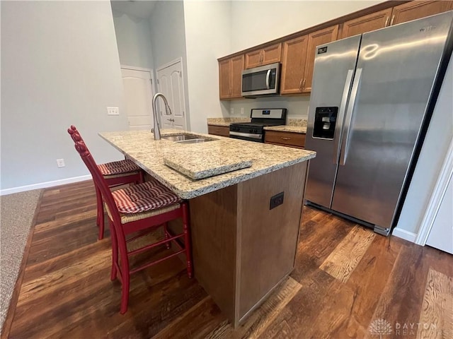 kitchen featuring dark wood finished floors, a center island with sink, light stone counters, appliances with stainless steel finishes, and a sink