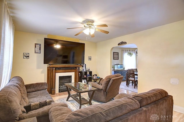 living area with baseboards, light wood-type flooring, a fireplace, arched walkways, and a ceiling fan