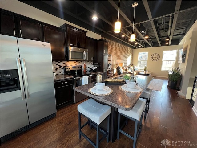 kitchen with dark wood-style floors, a center island with sink, a sink, appliances with stainless steel finishes, and a kitchen bar