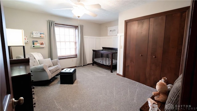 bedroom featuring a wainscoted wall, light carpet, a closet, a ceiling fan, and a nursery area