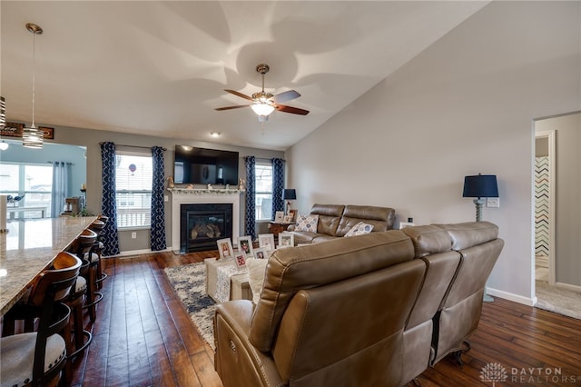 living area with lofted ceiling, a glass covered fireplace, dark wood-style floors, baseboards, and ceiling fan