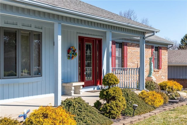 doorway to property featuring brick siding, roof with shingles, and covered porch