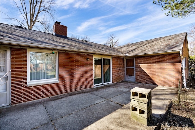 back of property with brick siding, a patio area, a chimney, and roof with shingles