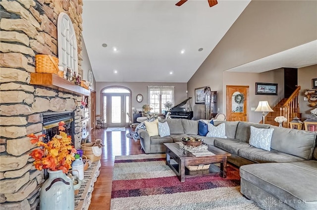 living area featuring stairway, a ceiling fan, wood finished floors, high vaulted ceiling, and a stone fireplace