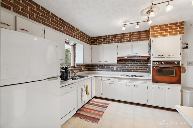 kitchen with brick wall, under cabinet range hood, light countertops, white cabinets, and white appliances