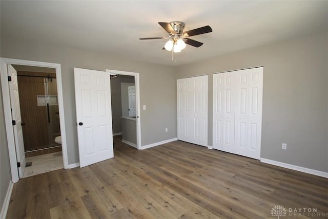 unfurnished bedroom featuring wood finished floors, baseboards, ceiling fan, ensuite bathroom, and two closets